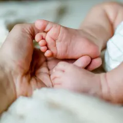 Dłoń dorosłego obejmuje stópkę niemowlaka na miękkim tle.