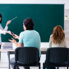 Nauczycielka z miarką w ręku zabiera telefon uczniowi przed tablicą.