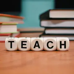 Kostki z literami "TEACH" na drewnianym stole, w tle stosy książek.