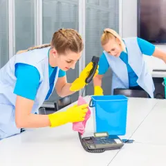 Dwie kobiety w żółtych rękawiczkach sprzątają biurko, jedna czyści telefon, druga zbiera śmieci.