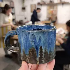 Ręcznie robiony, niebiesko-brązowy kubek ceramiczny trzymany w dłoni, w tle pracownia ceramiczna.
