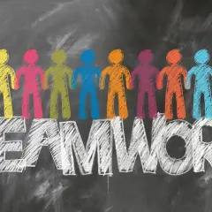 Kolorowe sylwetki ludzi stojące nad napisem TEAMWORK, narysowane kredą na tablicy.