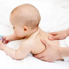 Niemowlę leżące na brzuchu, masowane przez dorosłego, trzymające zabawkę.