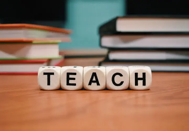 Kostki z literami "TEACH" na drewnianym stole, w tle stosy książek.