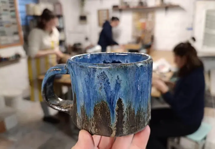 Ręcznie robiony, niebiesko-brązowy kubek ceramiczny trzymany w dłoni, w tle pracownia ceramiczna.