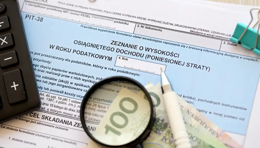 Formularz PIT-38, kalkulator, lupa, długopis i banknot 100 zł.