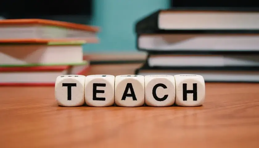 Kostki z literami "TEACH" na drewnianym stole, w tle stosy książek.