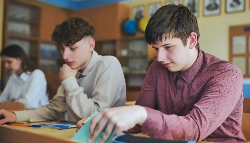 Uczniowie siedzą przy ławkach w klasie, jeden z nich przegląda notatki.