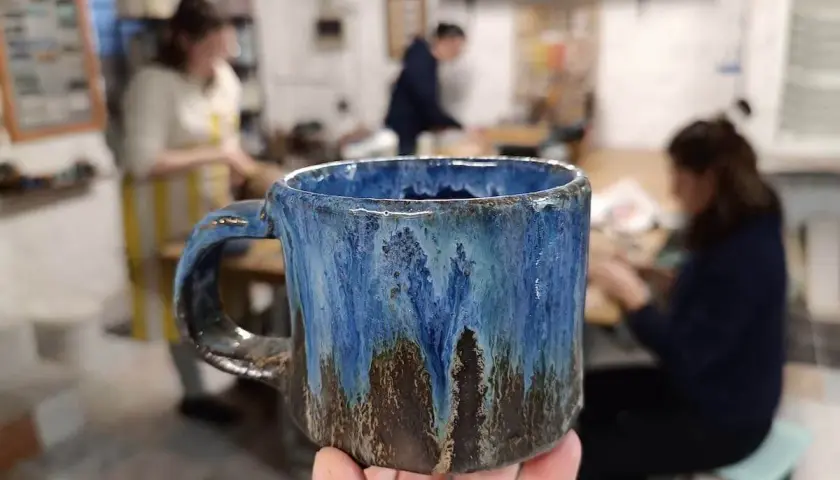 Ręcznie robiony, niebiesko-brązowy kubek ceramiczny trzymany w dłoni, w tle pracownia ceramiczna.