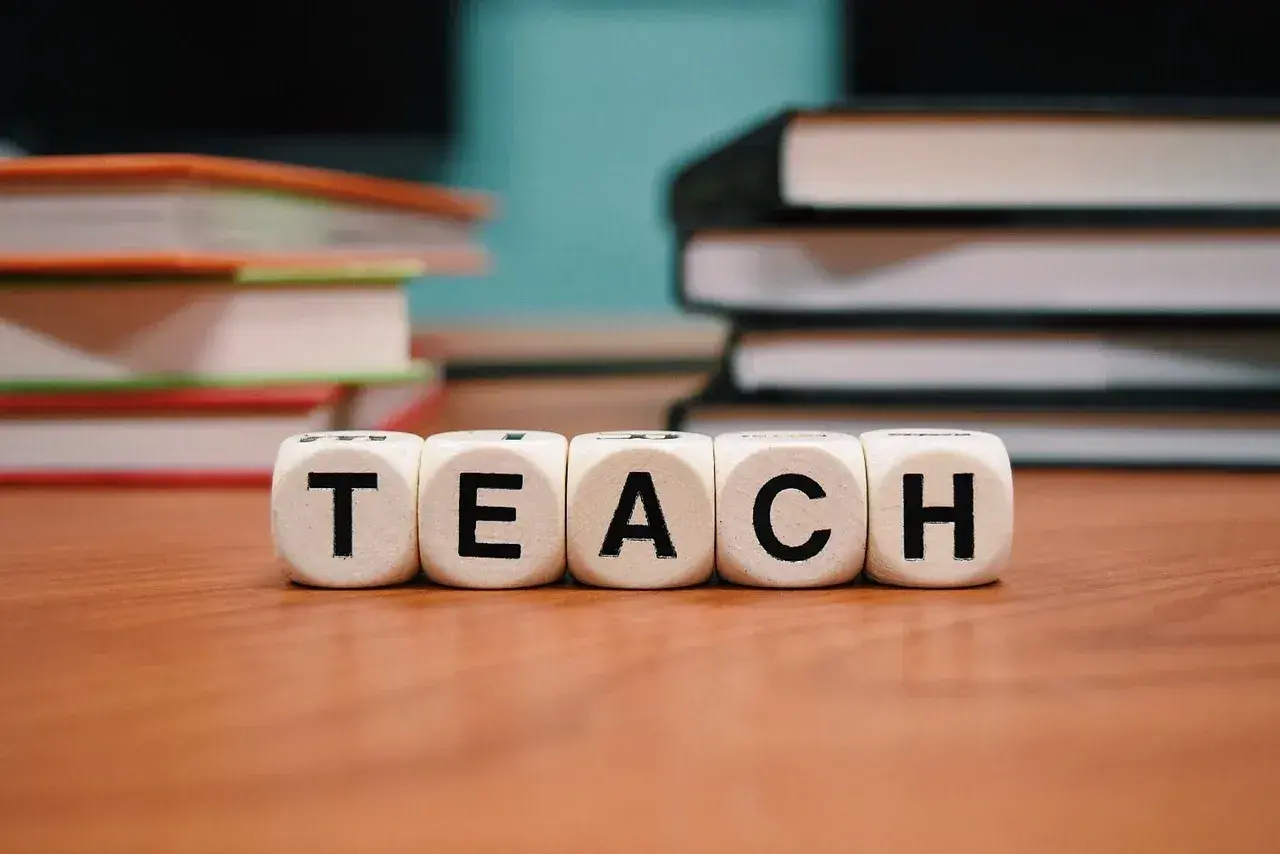 Kostki z literami "TEACH" na drewnianym stole, w tle stosy książek.