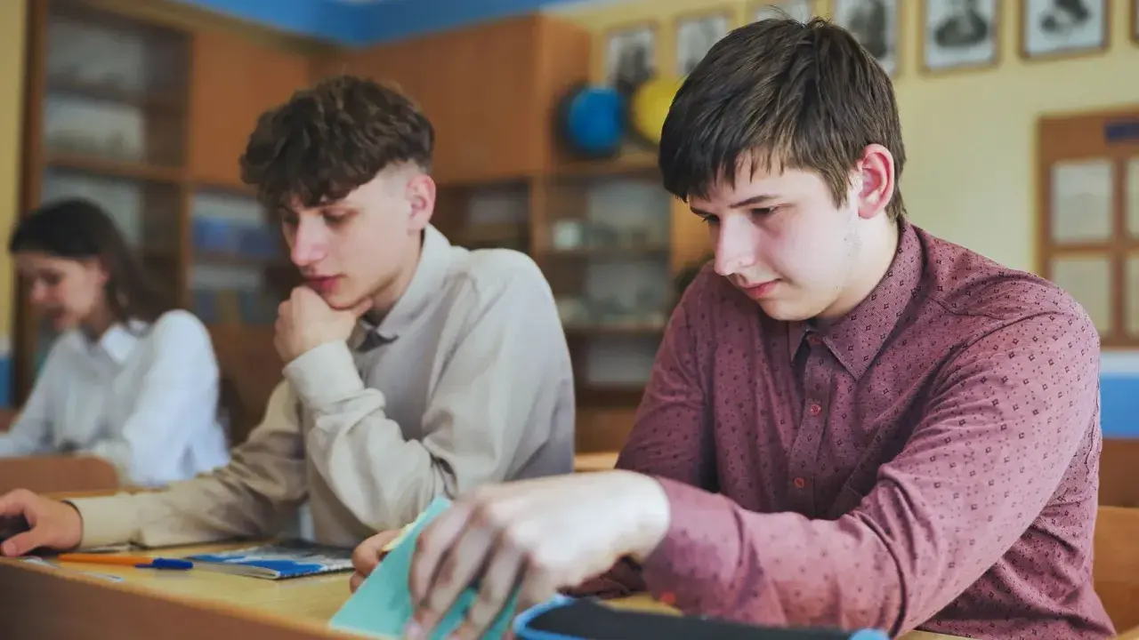 Uczniowie siedzą przy ławkach w klasie, jeden z nich przegląda notatki.