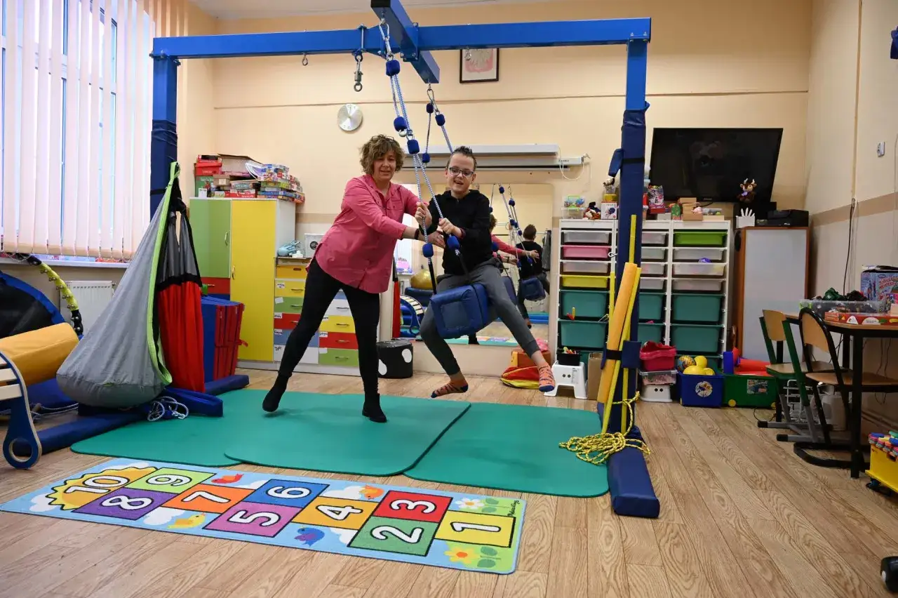 Kobieta i chłopiec ćwiczą na huśtawce terapeutycznej w sali integracji sensorycznej.